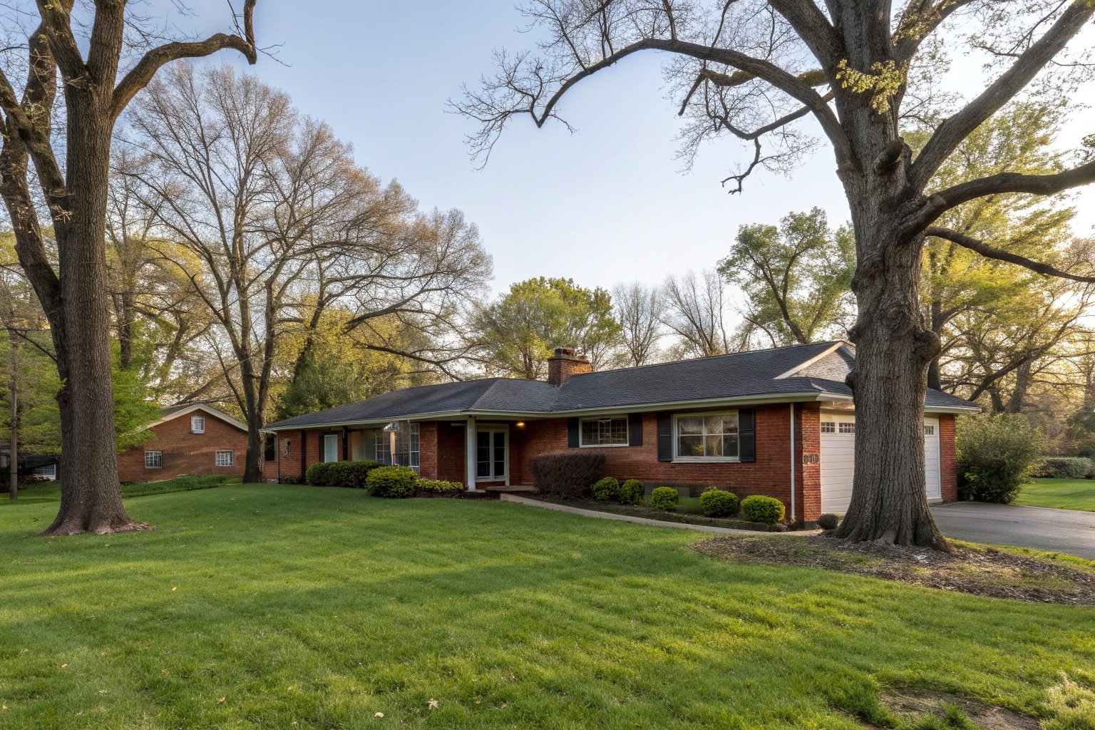 A single-story red brick ranch house with a charcoal shingle roof under big oak trees and a green lawn